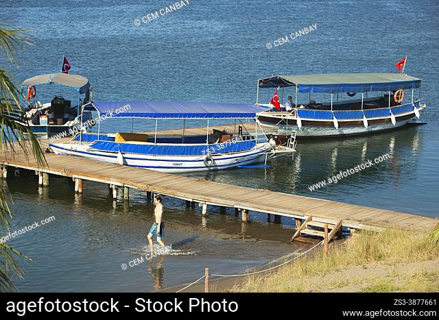 Touristic river boats near the wooden pier on Dalyan River at Iztuzu Beach-Turtle Beach with a young man entering into the water in the foreground