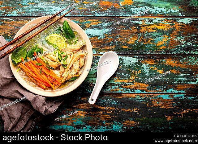 Asian soup with rice noodles, chicken and vegetables in ceramic bowl served with spoon and chopsticks on rustic wooden background from above with space for text