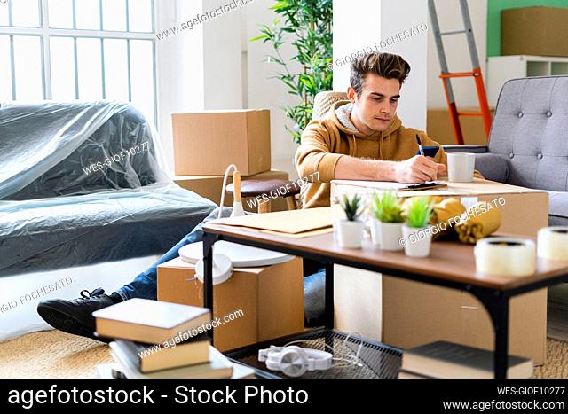 Young man writing on paper over cardboard box in new apartment