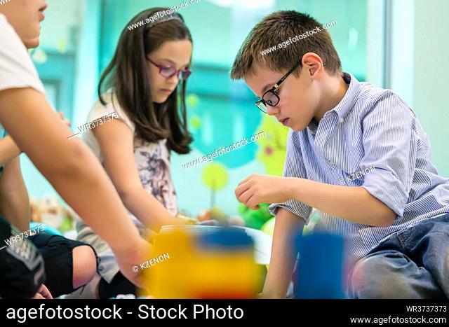 Cute girl wearing eyeglasses while playing with concentration a creative game next to her colleague in the classroom of a modern kindergarten