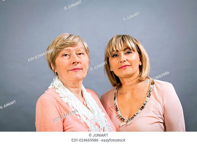 Attractive stylish blond senior lady with her beautiful middle-aged daughter posing together with her hands on her shoulders smiling at the camera on a grey...