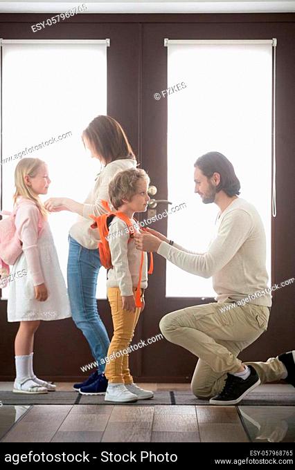 Young married couple and little children standing in hallway at home. Parents helps to preschool daughter and son gather to school or for a walk