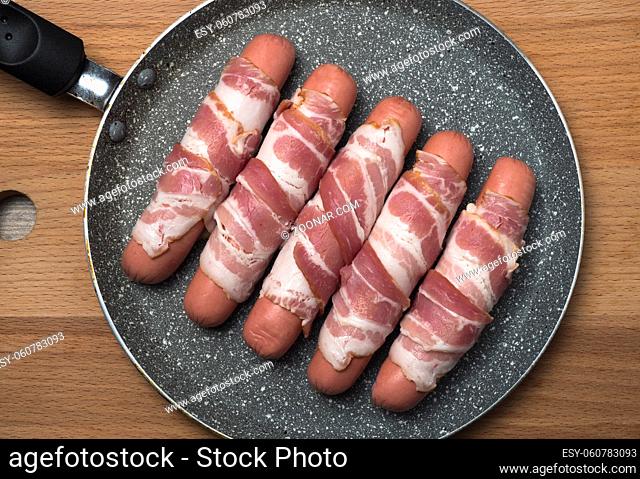 Preparation of raw sausages wrapped spirally in bacon on a frying pan. Top view