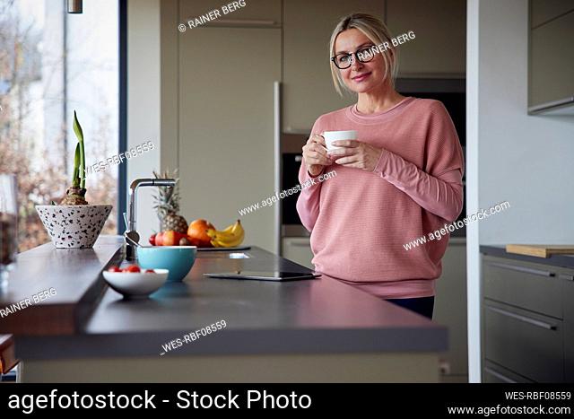 Blond woman wearing eyeglasses holding coffee cup standing in kitchen