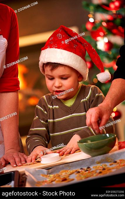 Small boy in santa claus hat helping at christmas baking watching mother and grandmother making cake