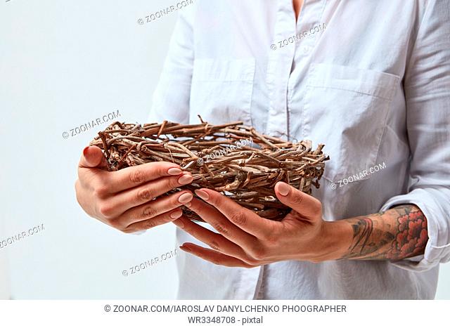 A woman with a tattoo is holding a nest of branches in her hands, against a gray background. Easter