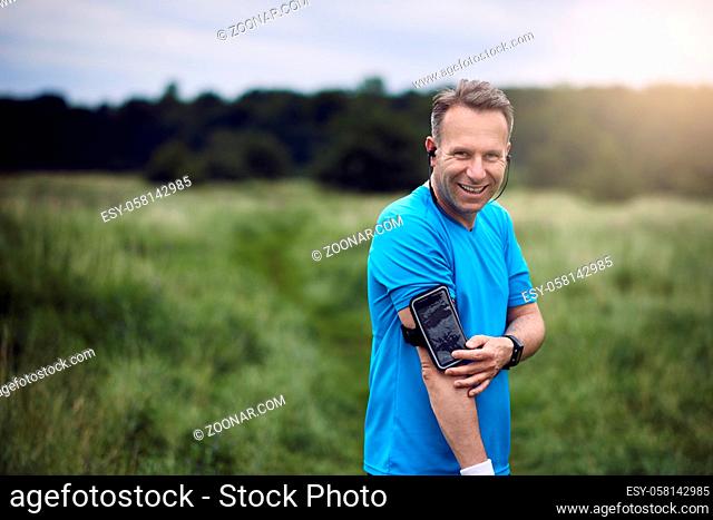 Cheerful mature man with smartphone on his arm listening to music while standing in meadow