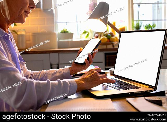 Senior caucasian woman sitting at table in kitchen, using smartphone and laptop with copy space