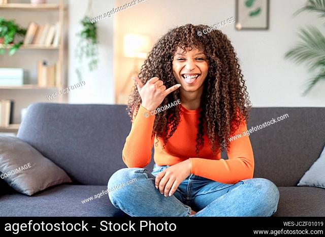Young woman gesturing shaka sign sitting on sofa at home