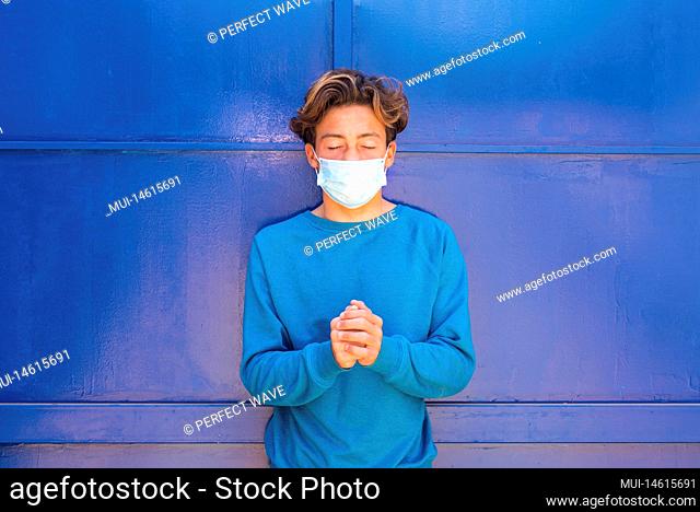 close up and portrait of teenager wearing a blue sweather with a blue background wearing a blue medical mask and praying alone to get off the virus coronavirus...