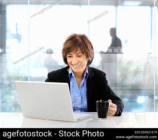 Happy senior businesswoman sitting at desk in modern corporate office, using laptop and drinking coffee