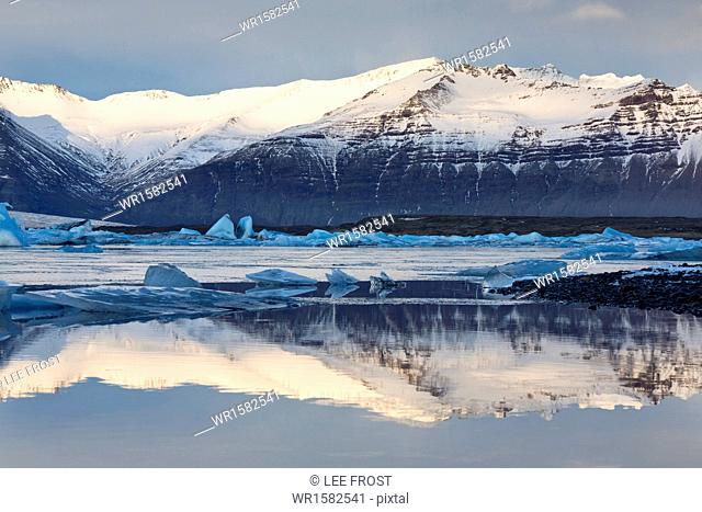 View over Jokulsarlon glacial lagoon towards snow-capped mountains and icebergs, with reflections in the calm water of the lagoon