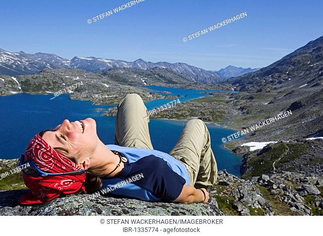 Young woman laughing, lying on a rock, relaxing, hiker enjoying the panorama on summit of historic Chilkoot Pass, Chilkoot Trail, Crater Lake behind