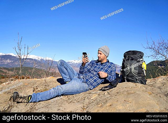Man using smart phone lying by backpack on sunny day