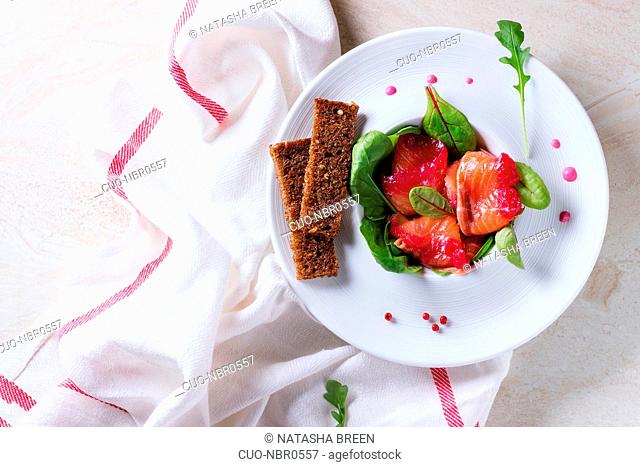 White ceramic plate with Sliced salmon filet, salted with beetroot juice, served with whole wheat toasts, salad leaves and beetroot sauce over white marble...
