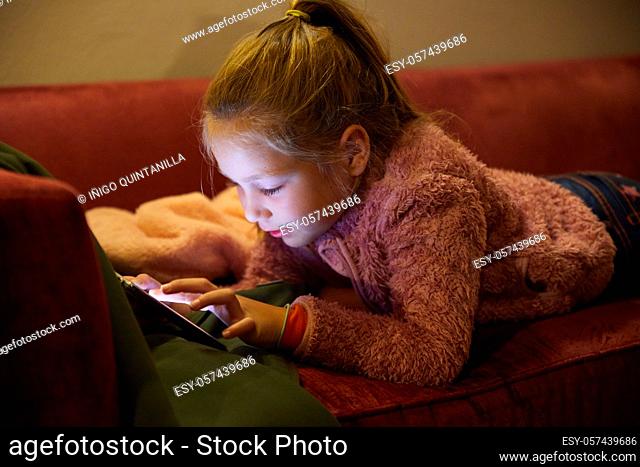 seven years old girl lying on red sofa at night touching digital screen mobile phone
