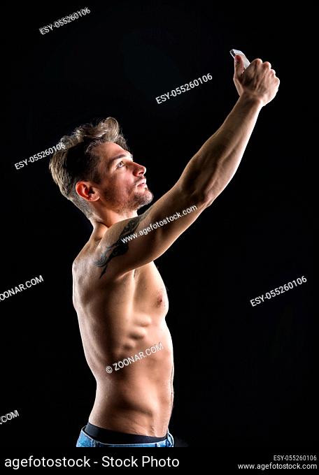 Handsome shirtless muscular young man using cell phone to take selfie picture, on dark background