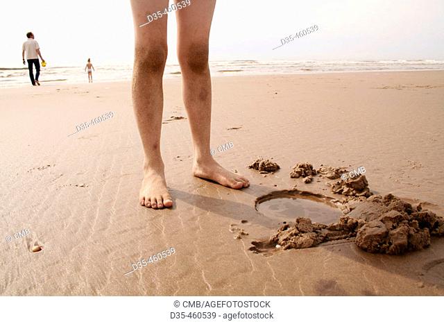 Kid's feet standing by water hole in sand, background: people on the beach