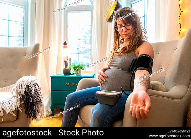 A beautiful young and healthy woman in later stages of pregnancy is seen monitoring blood pressure with a digital sphygmomanometer at home