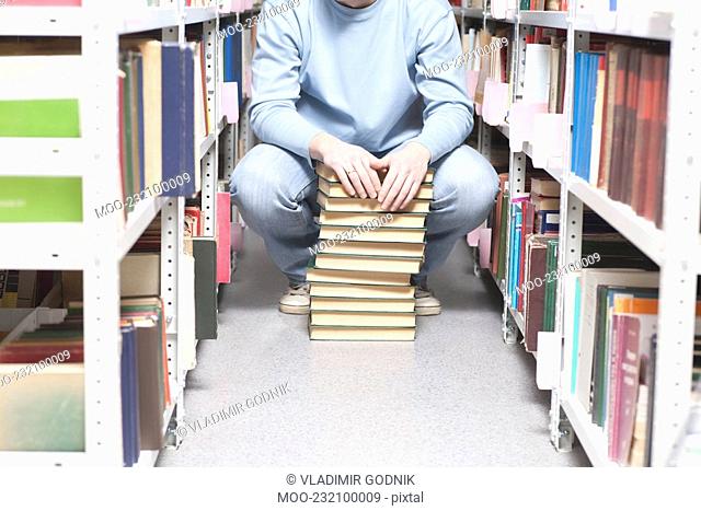 Young man crouches with pile of books in library