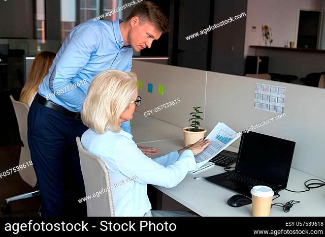 Serious company ceo mature businesswoman sitting at desk talking with employee male worker pointing on mistakes in financial report or sales statistics