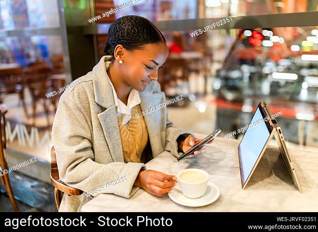 Teenage girl with coffee cup using smart phone by laptop at cafe