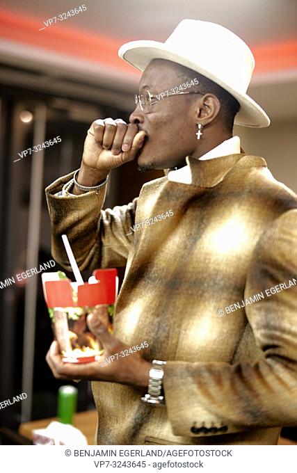 well-dressed stylish man choking while holding takeaway fast food doner kebab box with plastic fork, indoors, in Munich, Germany