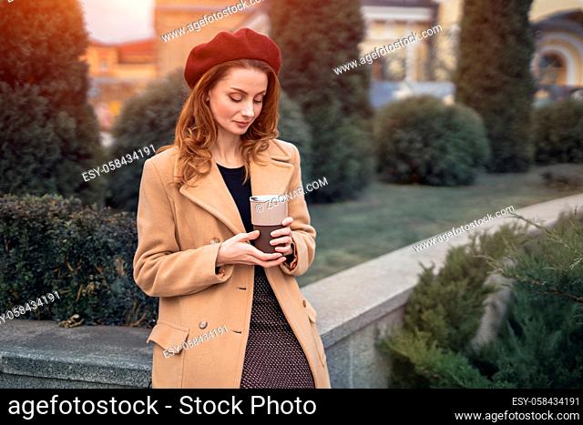 Portrait of stylish young woman wearing autumn coat and red beret outdoors. Pensive beautiful young woman holding a cup of coffee on the street female fashion