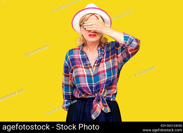 I don't want to see it. Portrait of scared or shocked modern stylish mature woman in casual style in white hat standing and closed her eyes with hand