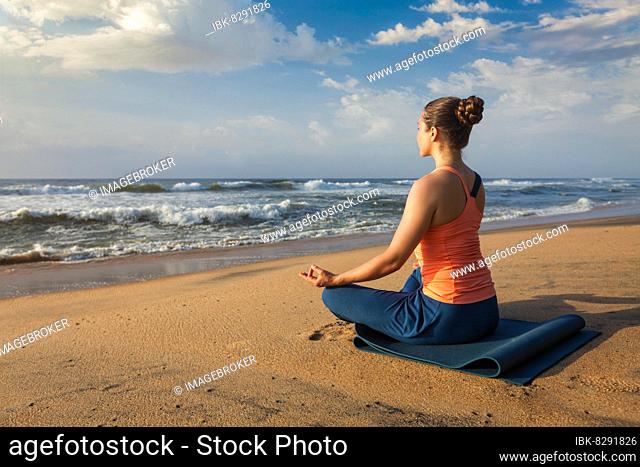 Woman doing yoga, meditating and relaxing in Padmasana Lotus Pose outdoors at tropical beach on sunset