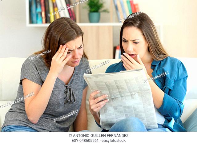 Worried friends reading newspaper news sitting on a couch in the living room at home