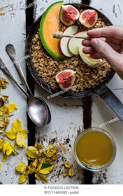 Honey being drizzled over roasted wholemeal oats in a cast-iron pan with fresh fruits