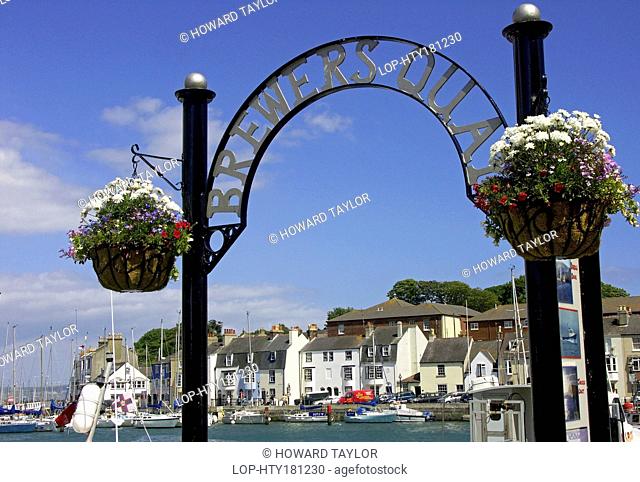 England, Dorset, Weymouth. Baskets of flowers hanging by a sign at the entrance into Brewers Quay in the seaside resort of Weymouth