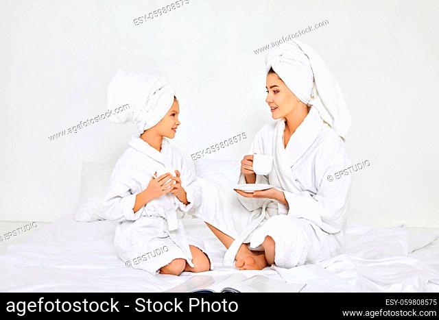 happy mother and daughter in white bathrobes and towels on heads drinking tea in bedroom