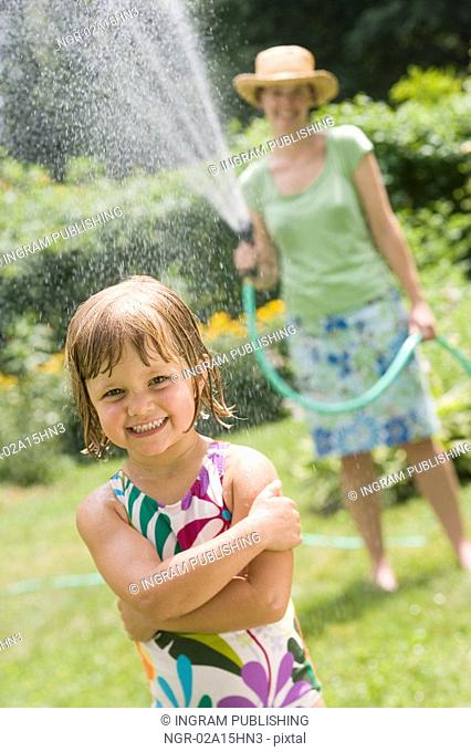 Pretty woman spraying little girl with water from a garden hose in the Summertime