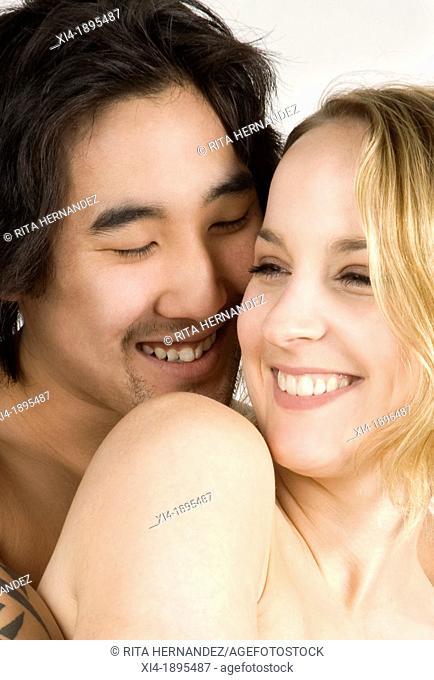 Close-up of Asian & caucasian couple hugging  He is close to her cheek with a smile and she is welcoming the hug  They both are showing bare shoulders  White...
