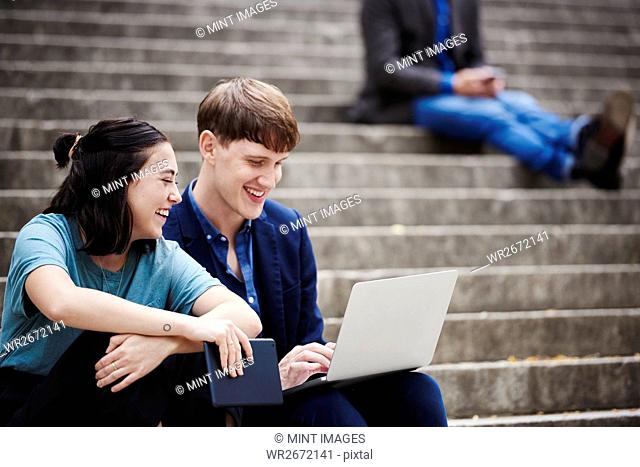 A young woman and a young man sitting on a flight of steps outdoors looking at a laptop together