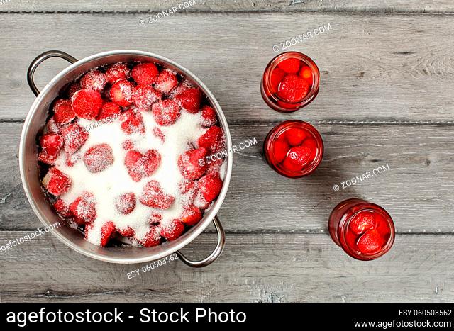 Tabletop view, steel pot with strawberries crystal sugar on top, three bottles with pickled fruit next to it. Homemade strawberry compote preparation
