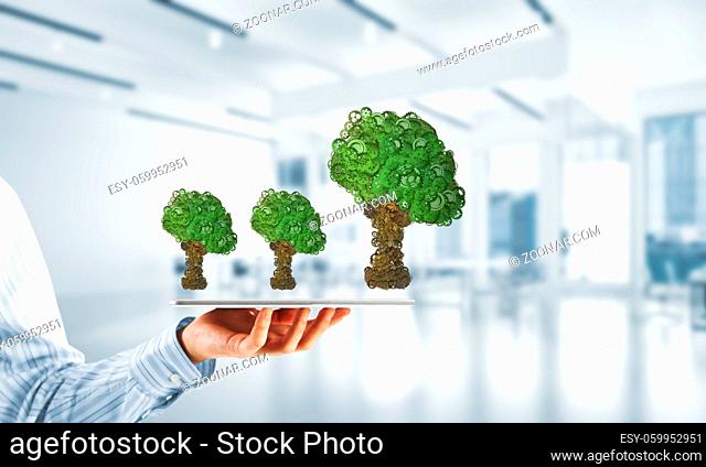 Close of businessman holding tablet pc with green tree made of gears and cogwheels. Mixed media