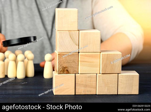 ladder of wooden cubes on a steel, behind a man under a magnifying glass examines wooden figures. Recruitment concept, search for talented employees