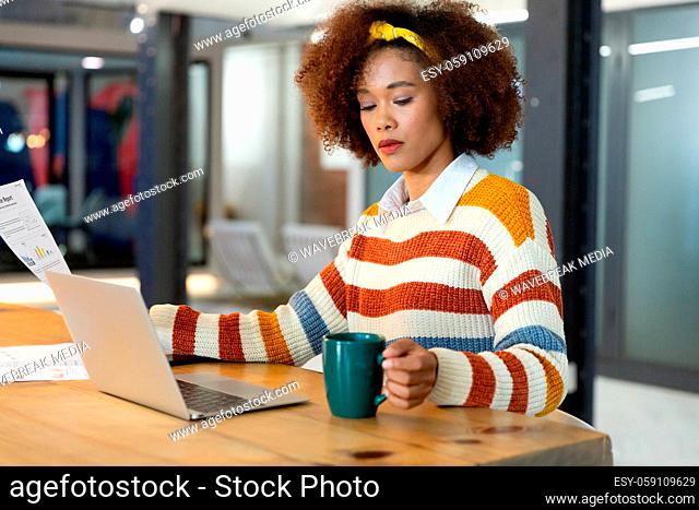 Portrait of mixed race businesswoman sitting at desk with laptop and holding coffee mug