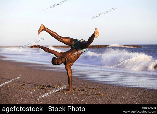 Shirtless African man doing handstand at beach against clear sky