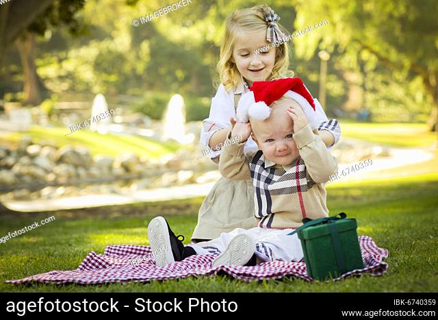 Sweet little girl tries to put A santa hat on her reluctant baby brother outdoors at the park