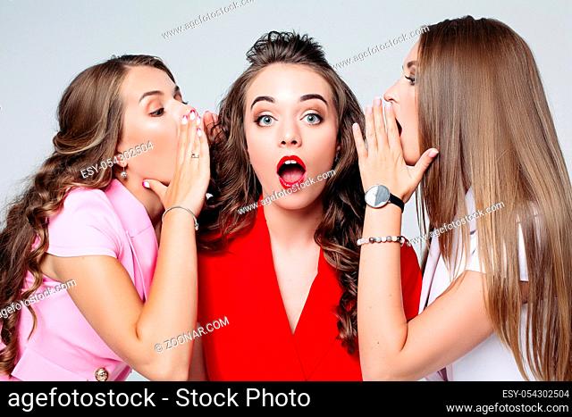 Portrait of shocked girl with long wavy brunette hair and red lips looking at camera with opened mouth while girlfriends whispering her secrets or gossiping