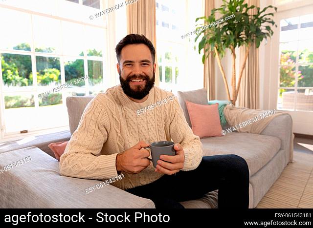 Portrait of happy caucasian man with beard smiling, sitting on couch in living room holding coffee