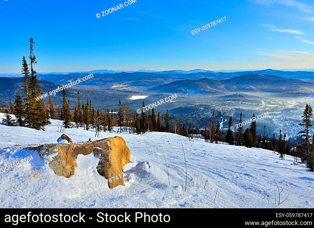 Panoramic winter landscape in the mountains of Western Siberia, Russia. Stone ledge on the snowy slopes, vast mountain ranges and valleys with pine forest...