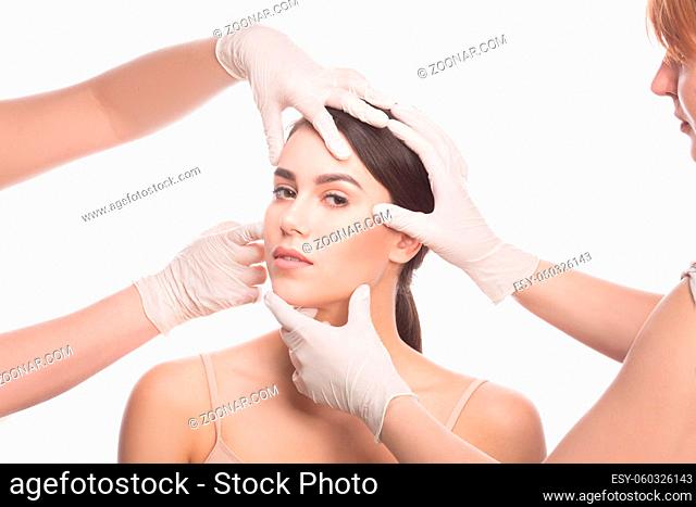 Handmade beauty. Beautiful young woman keeping eyes opened while four hands in medical gloves touching her face isolated on white background