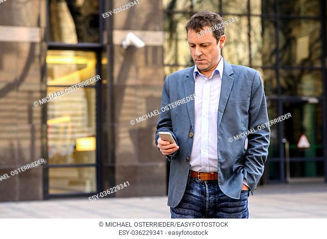 A middle age businessman standing in front of an office building while using his smartphone