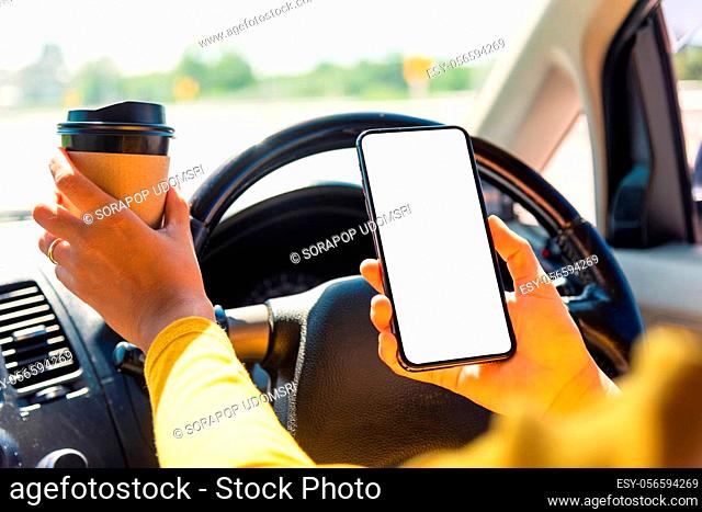 Asian woman drinking hot coffee takeaway cup inside a car and using smartphone blank screen while driving the car in the morning during going to work on highway