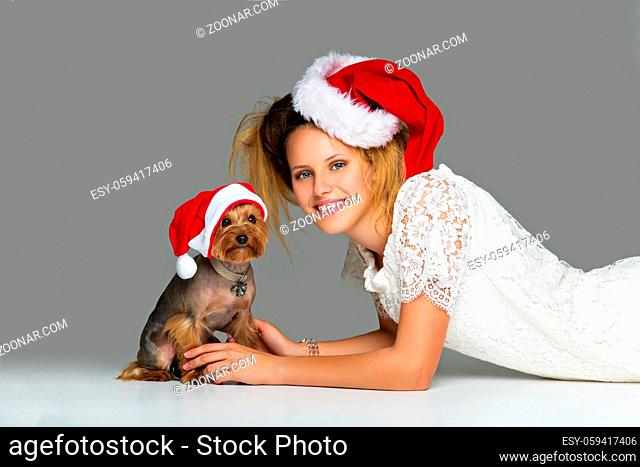 Beautiful girl in lace dress and red santa cap lying with pretty yourkshire terrier in christmas hat. Studio shot over grey background. Copy space